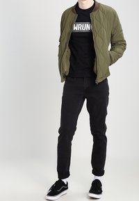 Hombre vestido con chaqueta acolchada verde oliva, camiseta gráfica negra, jeans negros arremangados, calcetines blancos y zapatillas negras, de pie contra un fondo liso.