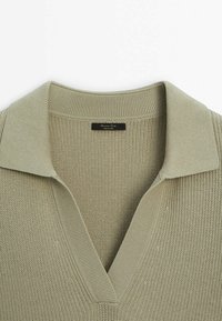 Pull polo en tricot vert olive clair. Présente un col en V et un col côtelé. La texture est respirante avec un motif fin. Étiquette visible.