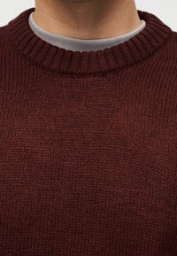 Homme portant un pull en maille rouge foncé par-dessus un t-shirt gris clair à col rond, mettant en valeur la texture détaillée de la maille et les couches de col.