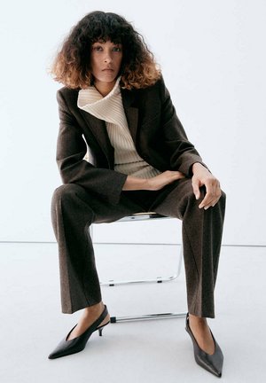 Femme aux cheveux bouclés assise sur une chaise, vêtue d’un tailleur marron, d’un pull côtelé crème et de talons slingback noirs à bout pointu.