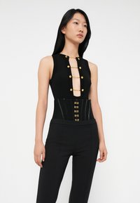 Woman wearing sleeveless black top with gold chains across deep neckline and a black corset belt with gold hooks, paired with black pants.