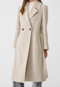 Cappotto doppiopetto in lana beige con ampio rever a incavo, tasche laterali e vestibilità sagomata, caratterizzato da una texture liscia e dettagli di cucitura discreti.