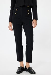Pantaloni neri a vita alta con una vestibilità slim, dotati di dettagli con bottoni dorati e una texture liscia. Abbinati a mocassini neri lucidi.