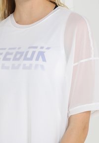 Vit tränings t-shirt med genomskinliga korta ärmar, tryckt logotyp i blått, rundad halsringning och avslappnad passform. Mjuk, lätt tyg.