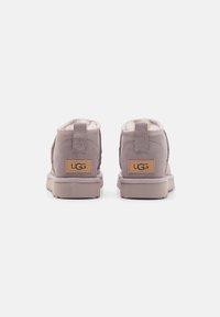 Botines de tobillo de ante morado con un forro suave y peludo, que presentan un parche de cuero marrón claro con el logo y una suela de goma plana.