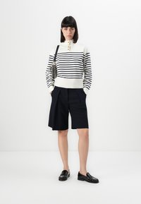 Maglione bianco lavorato a maglia con righe navy, caratterizzato da un collo alto e dettagli in bottoni, abbinato a pantaloni corti scuri su misura e mocassini neri.