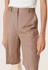 Donna che indossa pantaloni corti beige a vita alta, al ginocchio, con tasche laterali e una canotta bianca a costine infilata dentro.