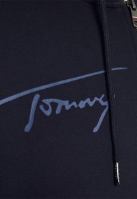 Donker marineblauwe stof met een blauw handgeschreven "Tommy" logo, een zwarte trekkoord met metalen punt, en een deel van een rits met een metalen trekker.