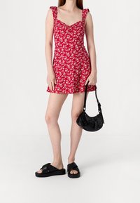 Punainen kukallinen lyhyt mekko neliökaula-aukolla ja röyhelöillä varustetuilla olkaimilla, yhdistettynä mustaan käsilaukkuun ja mustiin paksupohjaisiin sandaaleihin.