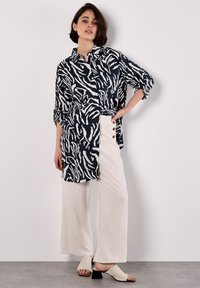 Una giovane donna posa contro un muro neutro, indossando una camicia a fantasia e pantaloni larghi chiari, abbinati a mule eleganti.