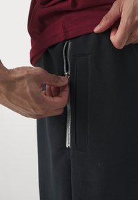 Calças de moletão pretas com um bolso lateral com fecho éclair. O tecido parece suave com uma textura mate. A peça de vestuário superior é bordeaux, parcialmente visível.