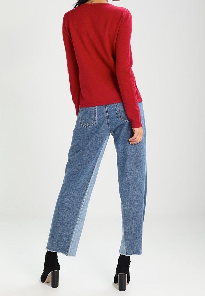 Personne portant un haut rouge à manches longues, un jean taille haute large bicolore, et des bottines noires à talons, debout devant un fond uni.