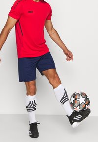 Hombre vestido con camiseta deportiva roja, pantalones cortos azul marino, calcetines blancos y botas negras, haciendo malabares con un balón de fútbol de color negro, blanco y naranja frente a un fondo liso.