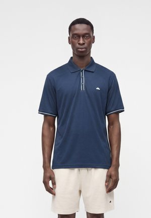 Homme portant un polo bleu marine à manches courtes et un short beige clair, debout sur un fond uni.