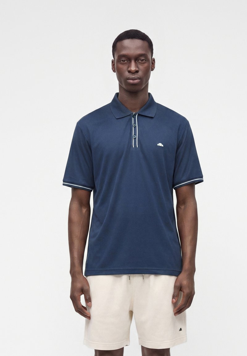 Homme portant un polo bleu marine à manches courtes et un short beige clair, debout sur un fond uni.