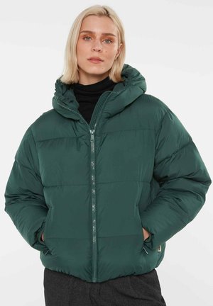 Frau mit kurzen blonden Haaren, die eine dunkelgrüne wattierte Winterjacke mit Kapuze, ein schwarzes Top und dunkelgraue Hosen trägt, Hände in den Taschen.