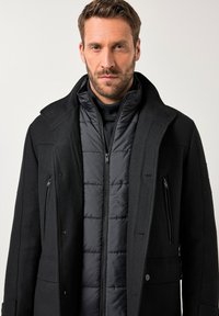 Uomo con capelli corti castani e barba, che indossa un giubbotto a strati nero imbottito e un cappotto di lana, con sguardo rivolto dritto davanti a sé e un'espressione neutra.