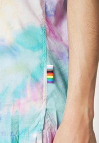 Maglietta tie-dye con colori pastello di turchese, rosa e viola. Include un'etichetta a strisce arcobaleno sul lato.