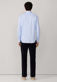Camisa de manga larga azul claro con dobladillo curvado, puños abotonados y un cuello clásico, combinada con pantalones oscuros navy y zapatos beige.