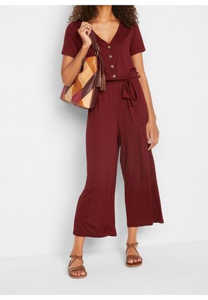 Donna che indossa una tuta bordeaux con bottoni e cintura in vita, che tiene una borsa in pelle multicolore e indossa sandali marroni con cinturini.