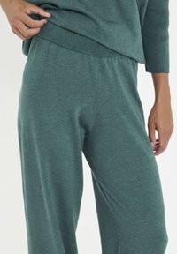Leichte grüne Strick-Loungewear-Hosen mit lockerem Schnitt, elastischem Bund und weicher Textur. Abgebildet mit einer Hand, die auf dem Bund ruht.