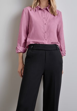 Kvinde iført en langærmet pink skjorte med knapper og opsmøgede manchetter, kombineret med højtaljede sorte bukser med knappedetaljer.