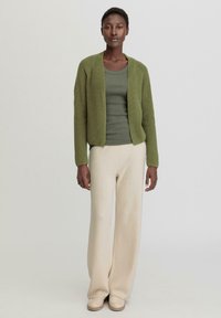 Grüner, gestrickter Cardigan über einem dunkelgrünen Oberteil, kombiniert mit off-white, weit geschnittenen Hosen und beigen Slippern. Einfaches, strukturiertes Design.