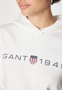 Gros plan d'une personne portant un sweat à capuche blanc avec le texte "GANT 1949" et un logo écusson rouge, blanc et bleu sur la poitrine.