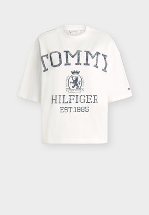 T-shirt blanc oversize avec "TOMMY" en grandes lettres usées, un blason de lion, "HILFIGER" et "EST.1985" imprimés en bleu marine.