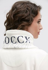 White fleece coat with a high collar, featuring dark blue lettering and light grey accents on the back. Smooth texture and casual design.