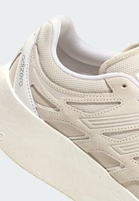 Ljusbeige träningsskor med ovandel i mesh och mocka, strukturerade mönster, vadderad krage och en vit gummisula. Varumärkeslogotyp synlig.