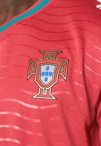 Κόκκινη αθλητική φανέλα με υφαντό σχέδιο, γιακά σε σχήμα V με γαλάζιες λεπτομέρειες και εμφανές κεντημένο εμβλημά της Πορτογαλίας.