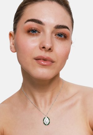 Collier en argent avec un cadre en perles vertes et un pendentif ovale présentant une image détaillée au centre. Design de chaîne simpliste.