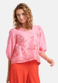 Rosa Bluse mit roter floralem Stickerei, V-Ausschnitt und dreiviertel Puffärmeln. Getragen mit strukturierten roten Hosen.