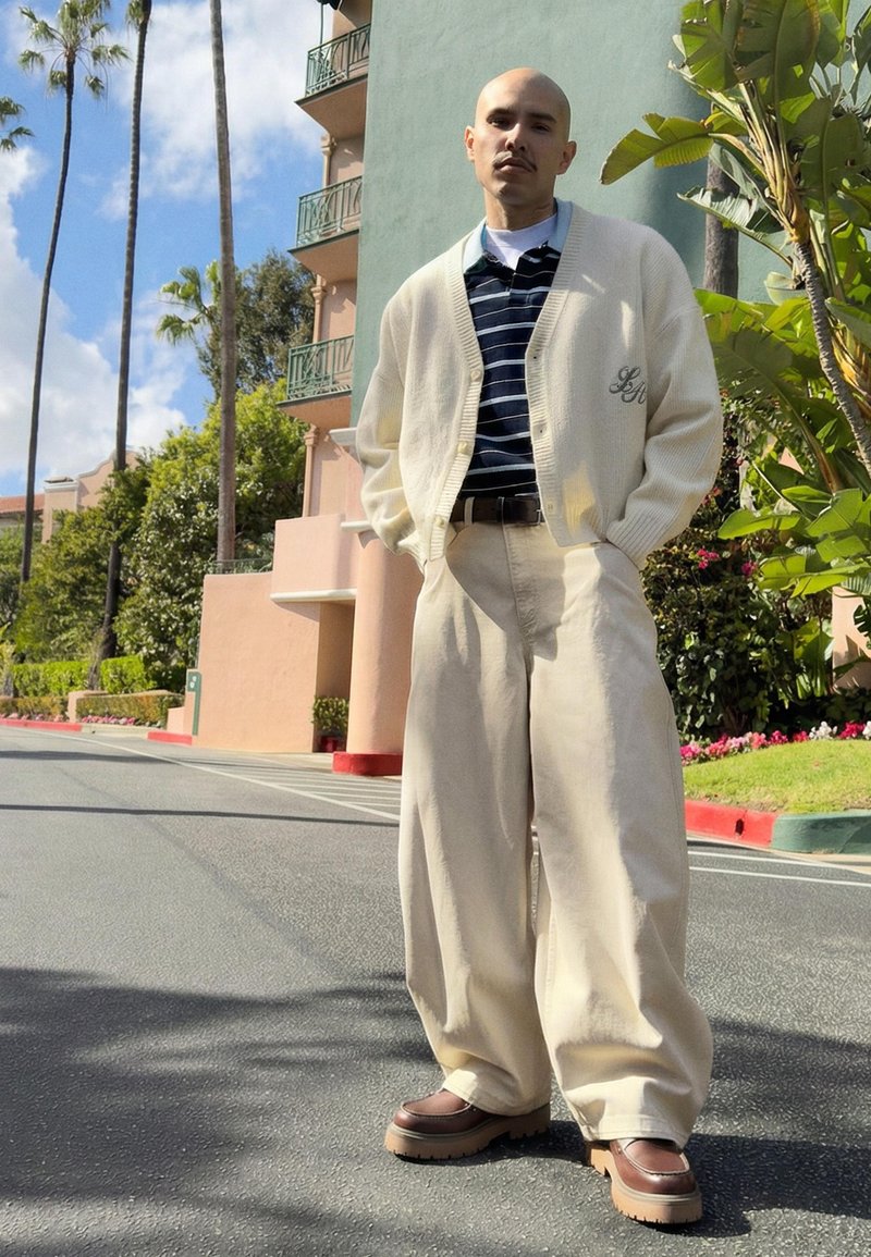 Plešatý muž v bílém kardiganu, pruhovaném tričku, širokých béžových kalhotách a hnědých botách stojí na slunné ulici poblíž palem a budovy.