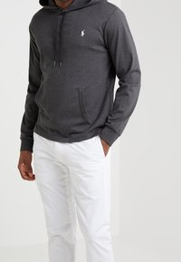 Grijze hoodie van zachte stof met een buidelzak aan de voorkant en een capuchon met trekkoord. Heeft een klein wit logo op de borst. Draag het met witte broek.