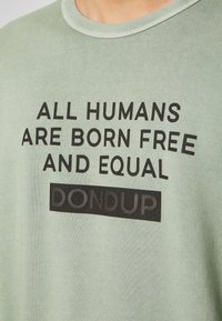 Ljusgrön bomull t-shirt med rund halsringning. Framsidan har fet svart text: "ALLA MÄNNISKOR KURSAS FRIA OCH LIKA" och "DONDUP."