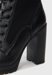 Black high-heeled ankle boot featuring a textured leather upper and canvas material, with lace-up design and thick, sturdy heel.
