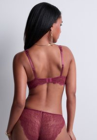 Femme aux longs cheveux noirs portant un ensemble de lingerie en dentelle bordeaux assorti, se tenant de dos à l'appareil photo sur un fond blanc.