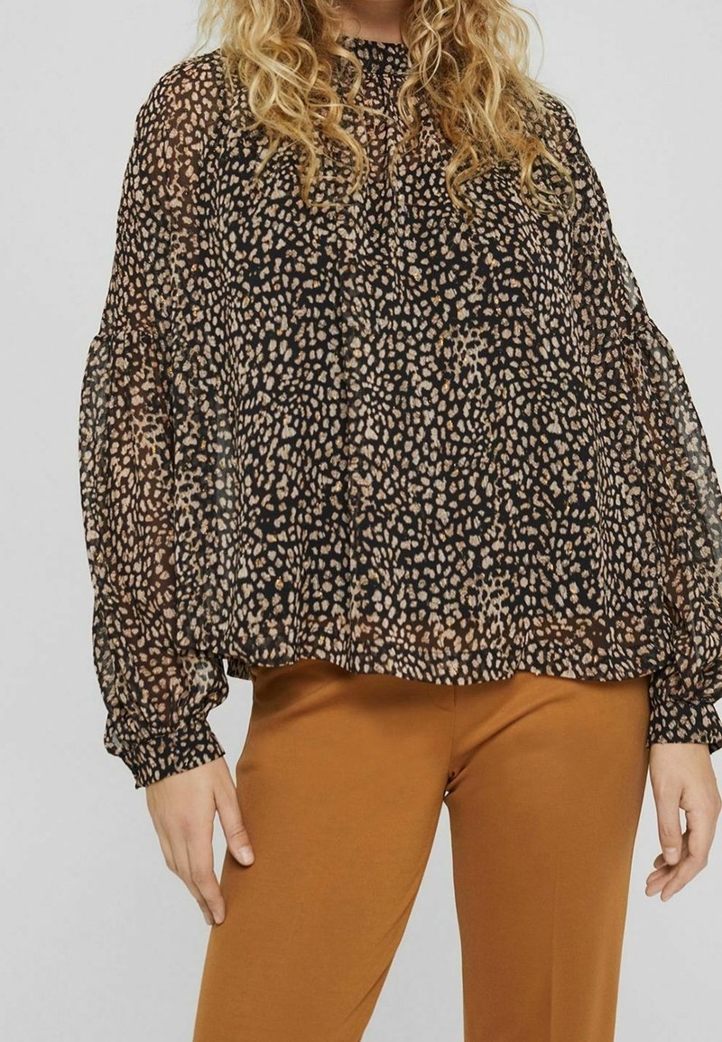Blusa nera trasparente con un motivo marrone chiaro e crema a macchie, con maniche arricciate e scollatura rotonda, indossata sopra pantaloni marroni.