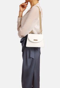 Borsa a tracolla in pelle bianca con pattina curva, tracolla in catena dorata e targhetta con logo. Indossata sulla spalla, abbinata a una blusa chiara e una gonna scura.
