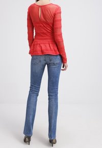 Top peplum rojo con mangas largas transparentes y detalle de abertura en la parte trasera, combinado con jeans ajustados azules y zapatos de tacón alto.