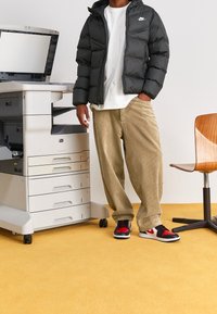 Black puffer jacket, white t-shirt, beige corduroy pants, and black/red sneakers. Standing next to a printer on yellow carpet.