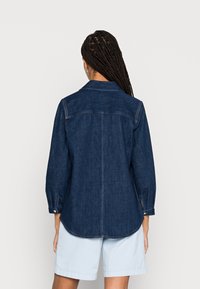 Camicia di denim, blu scuro, maniche lunghe con polsini a bottone e colletto a punta. Presenta un orlo curvo e cuciture a contrasto.