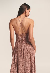 Rožinės su gėlėmis raštuoto suknelės nugaros vaizdas, su plonomis susikertančiomis petnešėlėmis ir surišimu nugarinėje dalyje, pagaminta iš lengvos, tekstūruotos medžiagos.