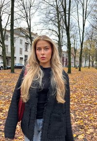 Giacca nera con trama testurizzata e bordi sfrangiati, indossata sopra un top nero. Lunghi capelli biondi ondulati, in piedi in un parco con foglie autunnali.