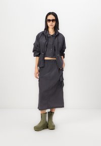 Slim-fit, cropped mørkegrå jakke med justerbare snorer, parret med et matchende langt nederdel og grønne ankelstøvler. Brune solbriller fuldender looket.