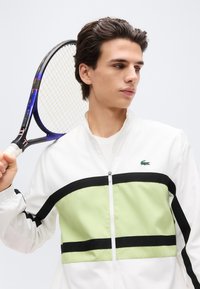 Νεαρός άνδρας με λευκό και ανοιχτό πράσινο μπουφάν κρατά ρακέτα του τένις στον ώμο του, κοιτώντας προς το πλάι σε ένα απλό φόντο.