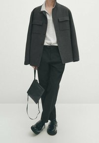 Zwarte wollen jas met twee borstzakken, lichtgrijs overhemd met kraag, zwarte broek en glanzende zwarte schoenen. Zwarte schoudertas inbegrepen.