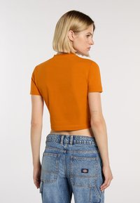 Camiseta corta naranja de manga corta, combinada con pantalones cargo de denim azul que presentan bolsillos y un parche con marca en la parte posterior.
